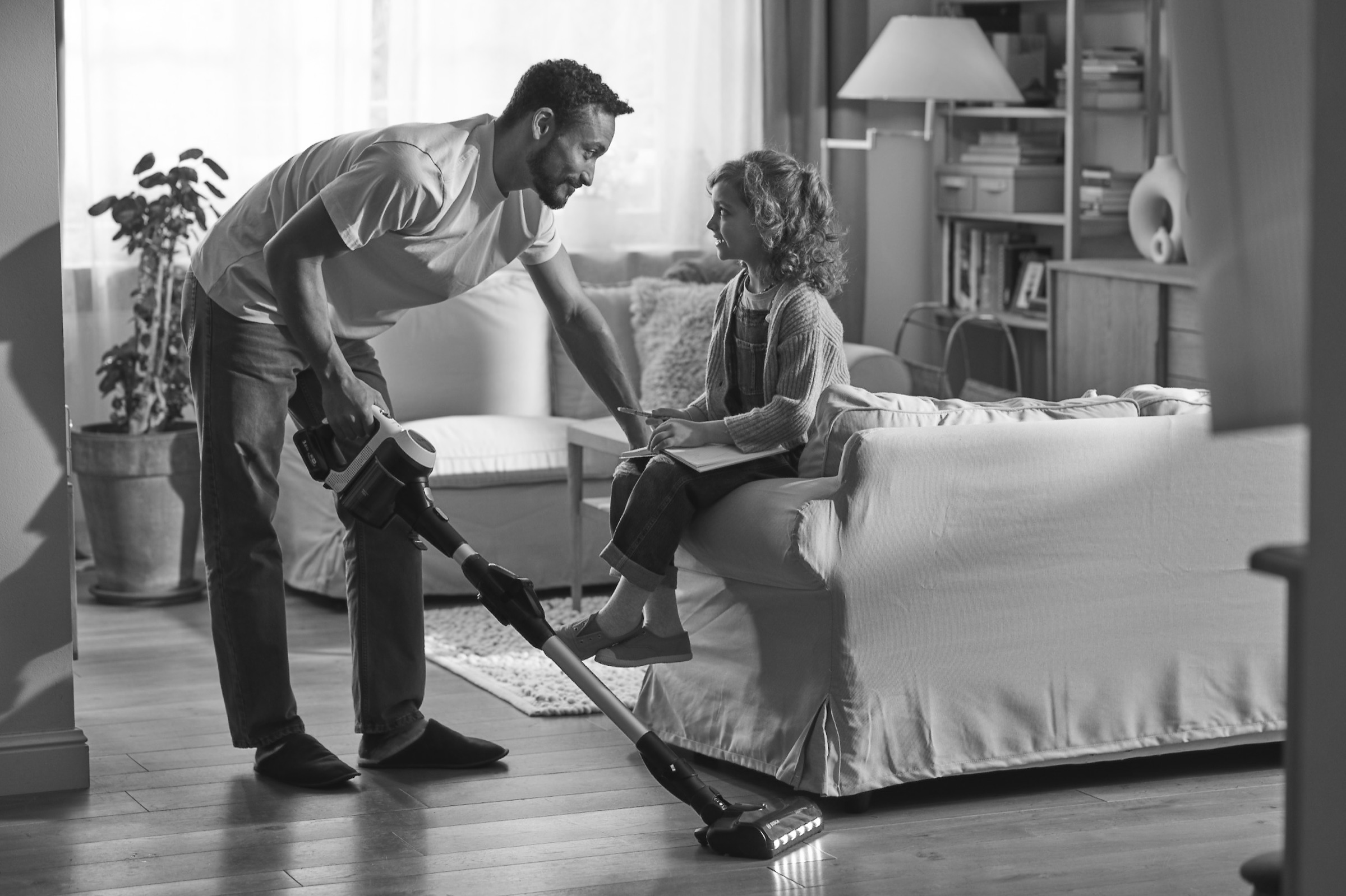 Father vacuuming while daughter sits on the sofa — a conversation mid-task