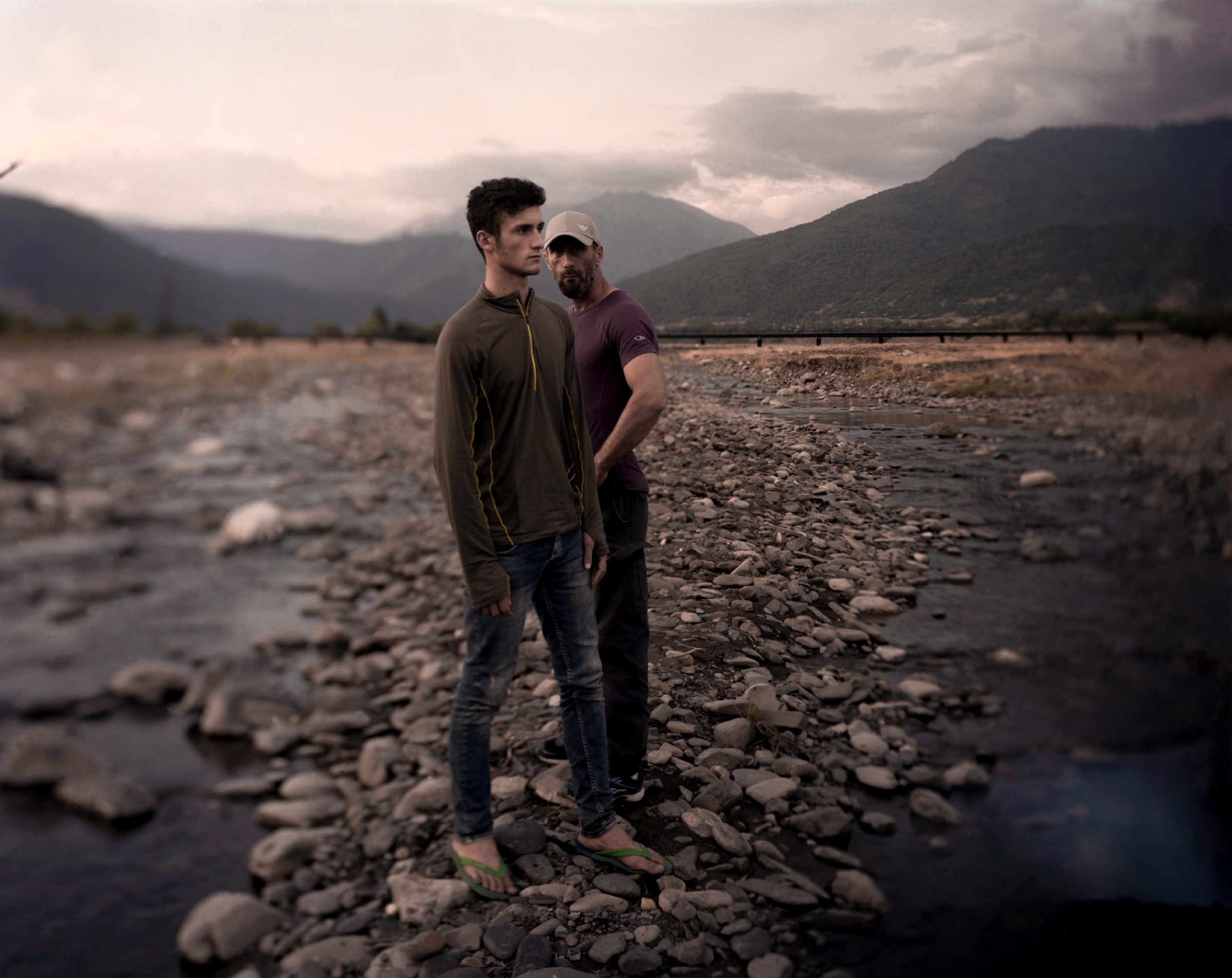 A father and son standing together on a rocky riverbed, mountains in the distance — Pankisi Valley