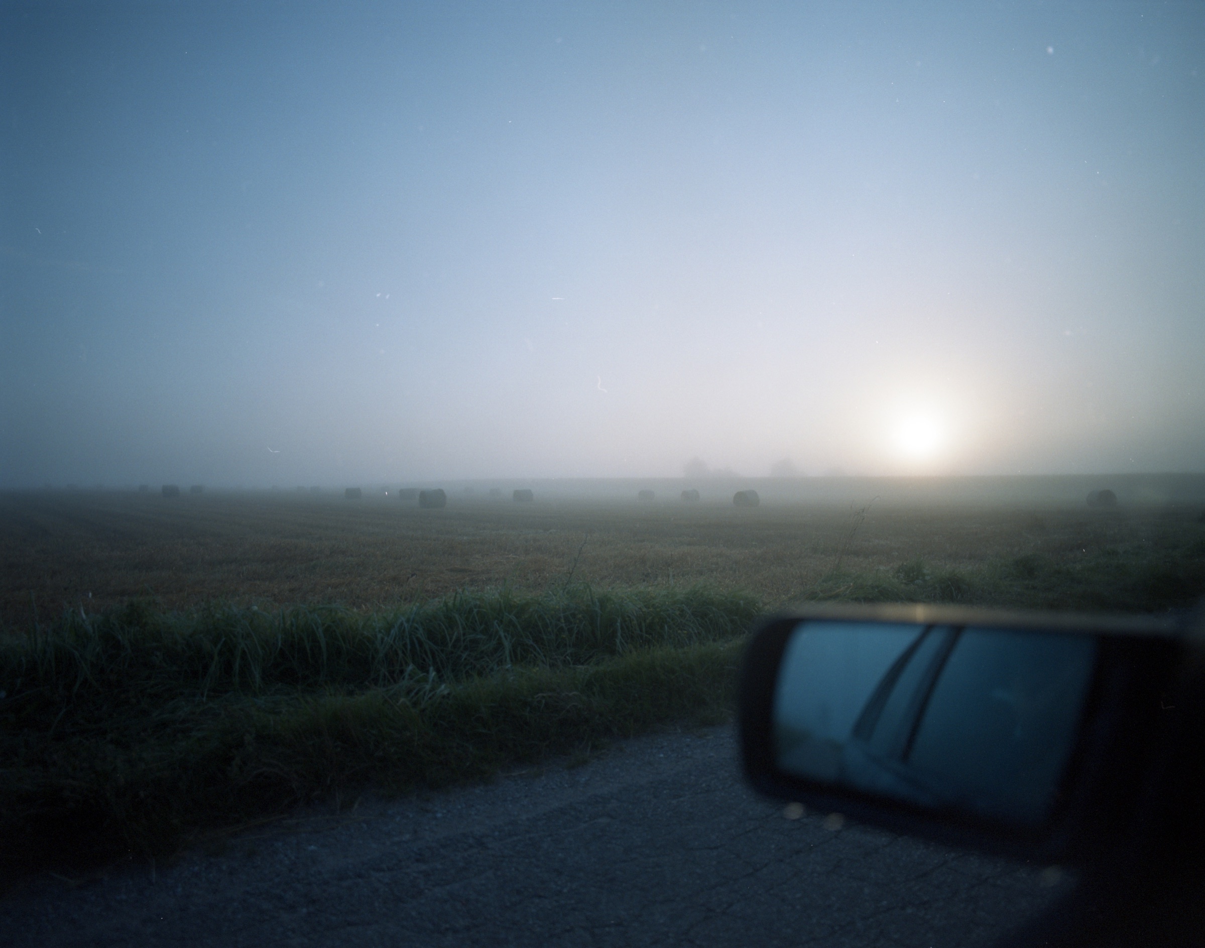 Foggy morning field, hay bales disappearing into mist, car side mirror in foreground — Podlasie