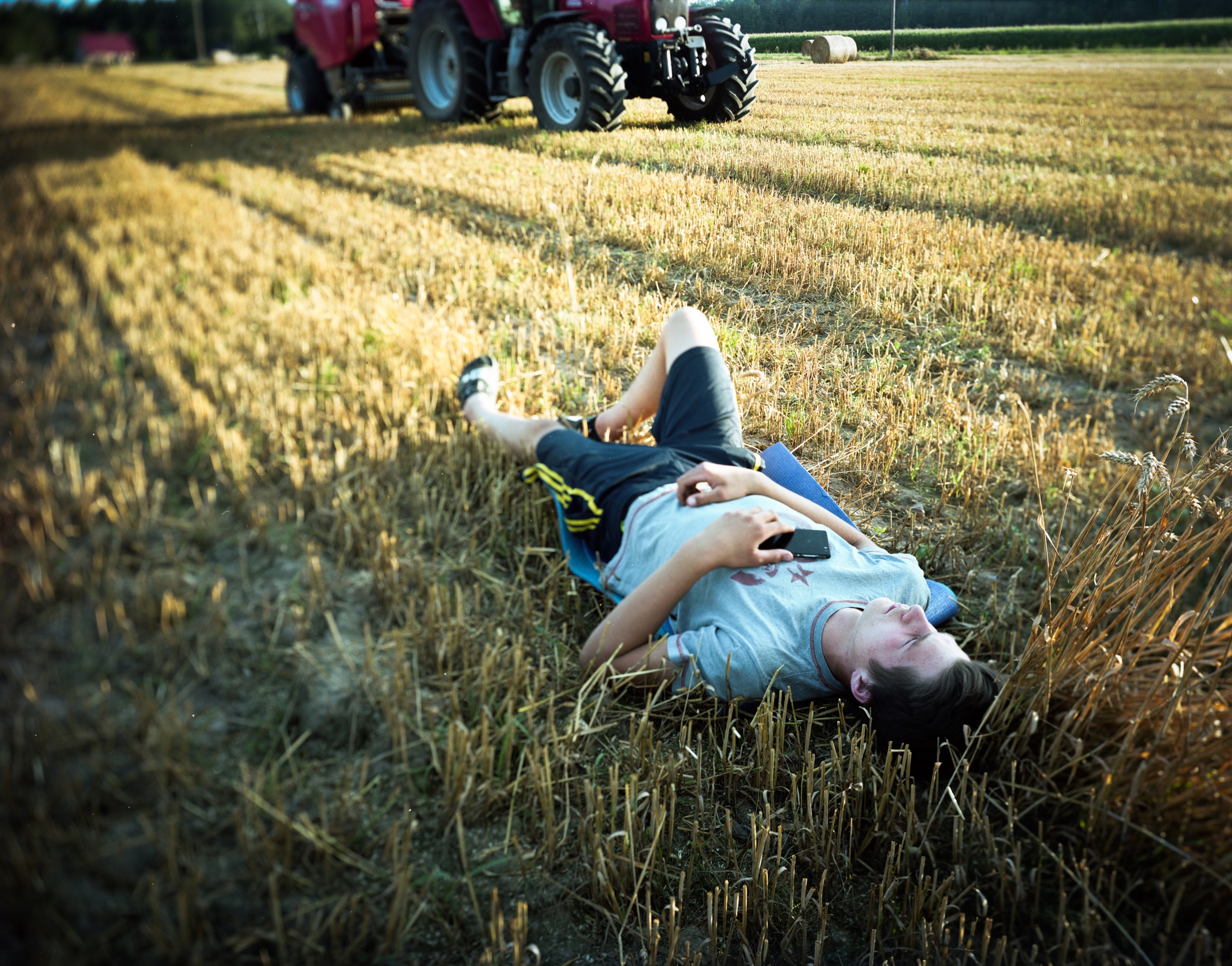 Young man lying on a stubble field with a phone, red tractor in the background — Podlasie