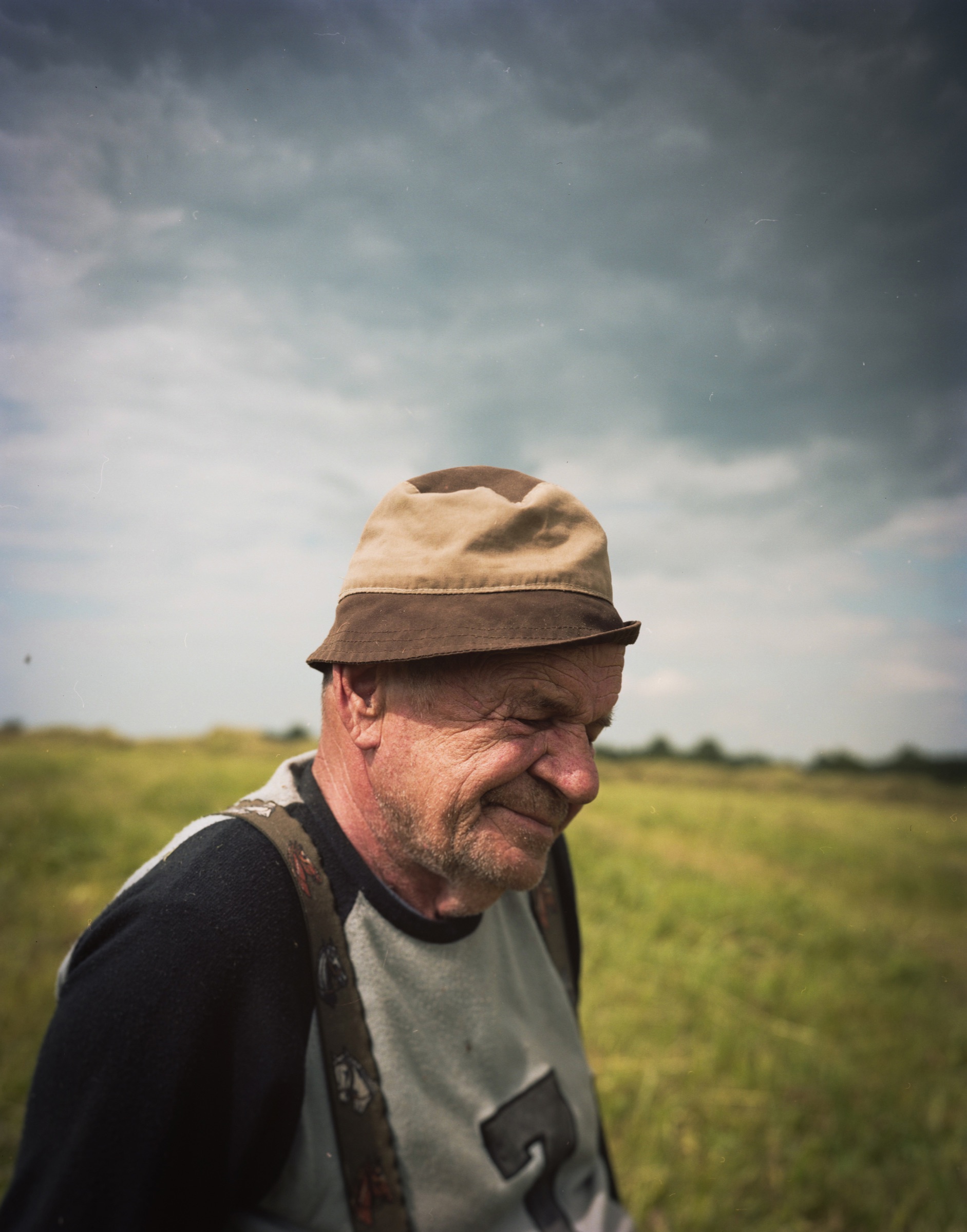 Older man in a hat, looking down, storm sky and open field behind him — Podlasie